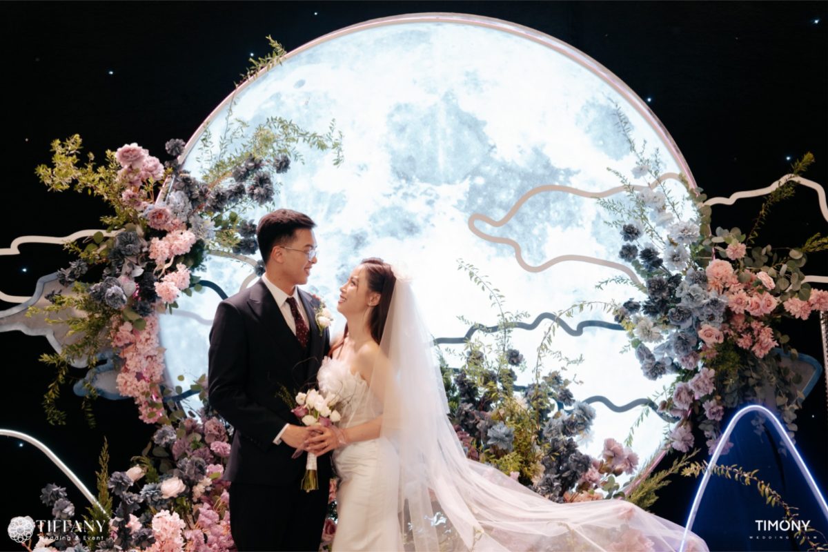 Cặp đôi chụp hình cưới trước sân khấu mặt trăng nghệ thuật, trang trí hoa tươi tông xanh tím lãng mạn. (Ảnh: TIFF.vn) 