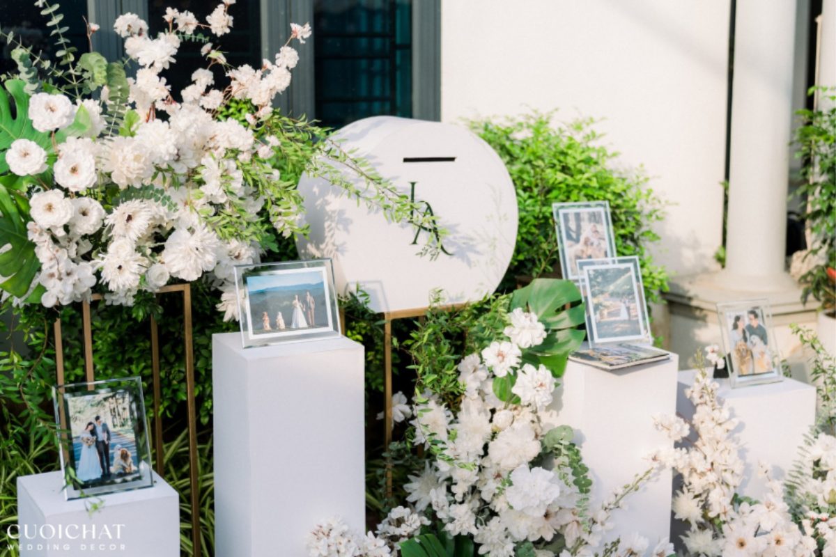 Bàn gallery lễ ăn hỏi ngoài trời trang trí hoa trắng, lá xanh và khung ảnh đôi trên bục trắng, mang vẻ thanh lịch và hiện đại. (Ảnh: TIFF.vn)