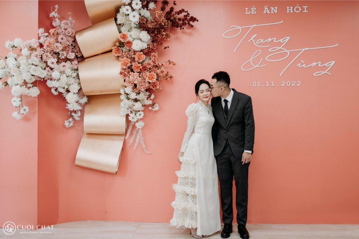 Chú rể hôn má cô dâu đầy ngọt ngào trước backdrop đỏ – cam rực rỡ, tạo khoảnh khắc lãng mạn và ấm áp trong ngày cưới. (Ảnh: TIFF.vn)
