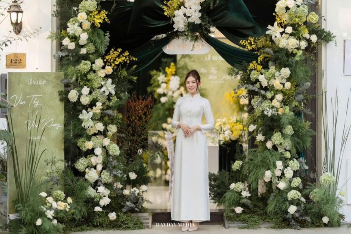 Cô dâu diện áo dài trắng duyên dáng bên cổng hoa trắng điểm lá xanh, tạo nên không gian lễ cưới tinh khôi và thanh nhã. (Ảnh: Sưu tầm)