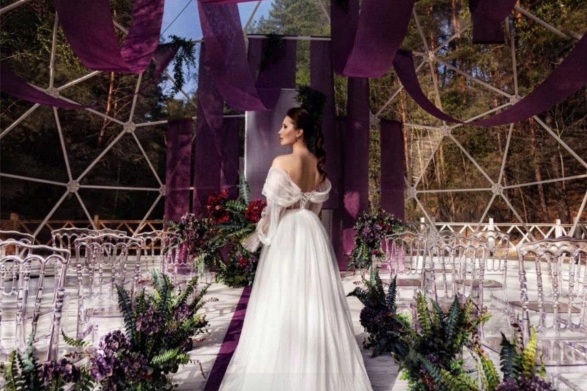 Cô dâu trong váy cưới trắng đứng trên lối đi tím lãng mạn, tiệc cưới ngoài trời với draping vải tím và hoa lá tươi, không gian dome hiện đại (Ảnh: Sưu tầm) 